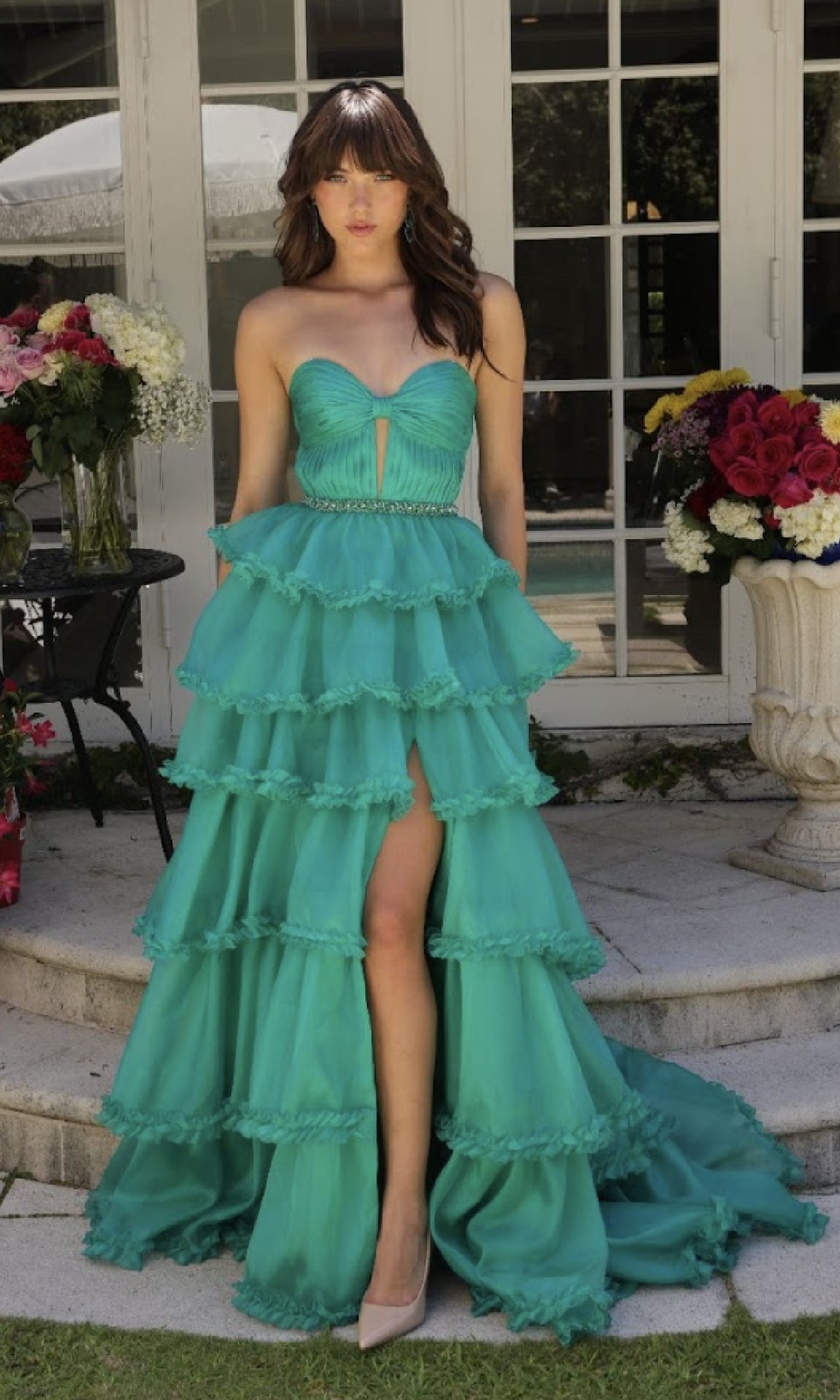 A woman stands on outdoor stone steps beside vases of red, pink, and white flowers, wearing the Ava Presley 29543 strapless prom ball gown with a sweetheart bodice, teal ruffled A-line skirt, and front slit.